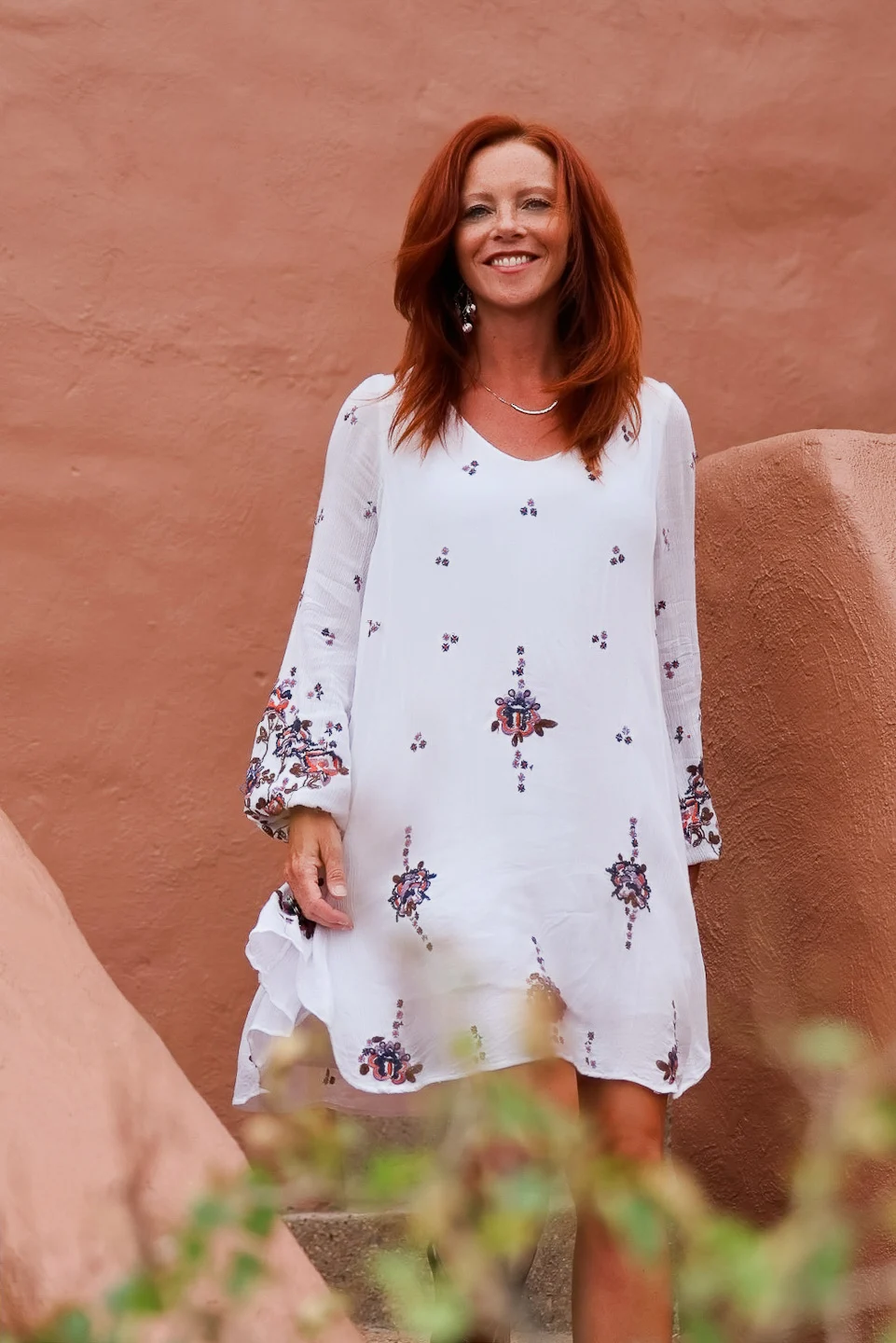 Angela smiling in a white embroidered dress on a stucco stairway with greenery in the foreground